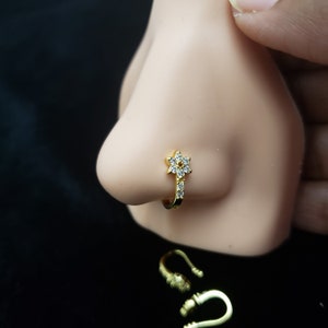 Puede incluir: Un aro de nariz dorado con un diseño floral con piedras claras y brillantes. El aro está unido a una banda curva y se muestra en una nariz de maniquí. El fondo es negro.