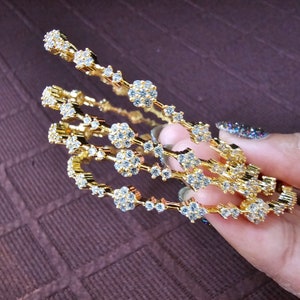 May include: Four gold bangles with clear cubic zirconia stones. The bangles are stacked on top of each other and are held in a hand.