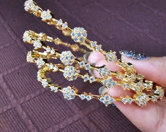 Puede incluir: Cuatro brazaletes de oro con piedras de circonita cúbica transparentes. Los brazaletes están apilados uno encima del otro y se sostienen en una mano.
