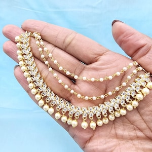 Puede incluir: Joyería dorada con cristales transparentes y perlas de imitación. La pieza presenta un diseño floral con perlas colgantes y delicadas cadenas. La joyería se sostiene en una mano sobre un fondo azul claro.