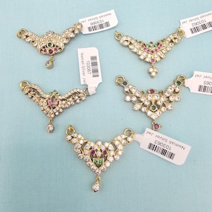 May include: Five gold-toned necklaces with intricate designs. Each necklace features a central pendant adorned with clear and colored gemstones, including red and green accents. Small tags with text are attached.