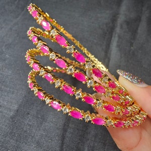 Pode incluir: Quatro pulseiras de ouro com pedras rosa e brancas. As pulseiras estão empilhadas umas sobre as outras e estão sendo seguradas por uma mão.