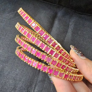 May include: Set of five gold bangles with pink square-shaped gemstones. The bangles are stacked on top of each other.