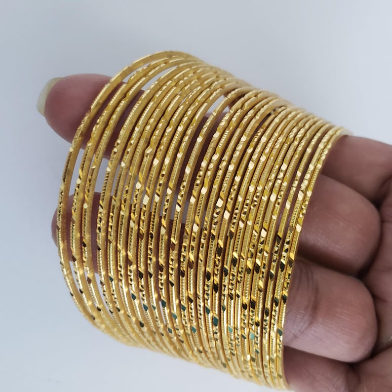 Może przedstawiać: Stos 24 złotych bransoletek z fakturowanym wzorem diamentowym. Bransoletki są cienkie i delikatne.