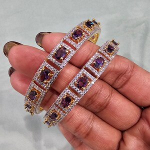 May include: Two gold bangles with a white and purple stone design. The stones are set in a square shape with a smaller round stone in the center.