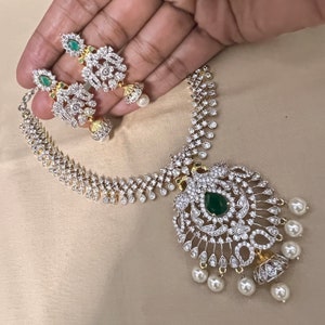 May include: A silver and gold necklace with a large, ornate pendant featuring a green gemstone surrounded by white stones. The pendant is accented with pearl drops. Matching earrings with green gemstones and white stones are also included.
