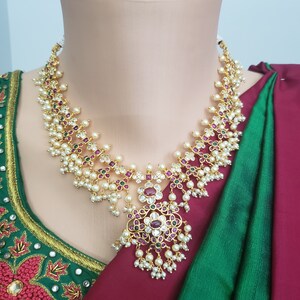 May include: A gold necklace with a pendant featuring a floral design with red, green, and white stones. The necklace is adorned with pearls.