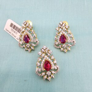 May include: A pair of silver earrings with a red stone in the center. The earrings are in the shape of a flower with a delicate design. The earrings are on a blue background.