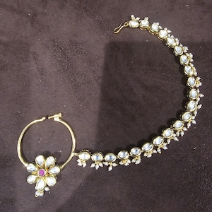Pode incluir: Um anel de nariz de ouro com um design floral e uma corrente de pequenas pedras brancas. A corrente está presa a um gancho.