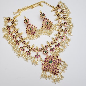 May include: A gold necklace and earrings set featuring pink and green stones with white pearl accents. The necklace has a large pendant with a floral design.