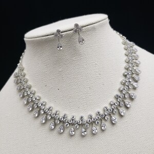 K&ouml;nnte beinhalten: Ein silbernes Halsketten- und Ohrring-Set mit klaren Zirkonia-Steinen. Die Halskette hat ein Tropfen-Design mit einer zarten Kette.