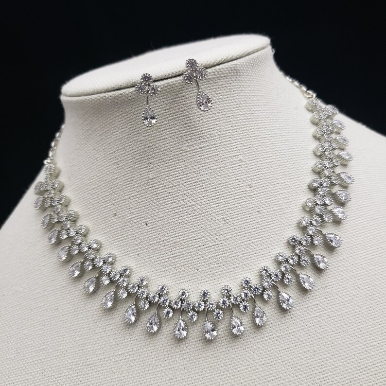 K&ouml;nnte beinhalten: Ein silbernes Halsketten- und Ohrring-Set mit klaren Zirkonia-Steinen. Die Halskette hat ein Tropfen-Design mit einer zarten Kette.