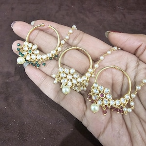 Pode incluir: Três argolas de nariz douradas com intrincados desenhos florais. Cada argola apresenta pedras preciosas claras e coloridas, juntamente com detalhes de pérolas penduradas. As argolas são conectadas por delicadas correntes de pérolas, mostrando um estilo de joalheria tradicional.