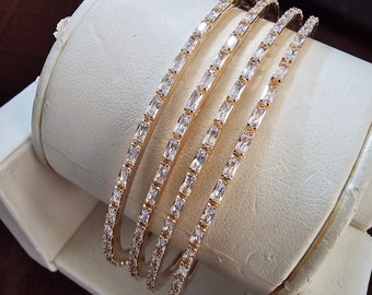 Puede incluir: Conjunto de cuatro brazaletes de oro con piedras de circonita cúbica transparentes. Los brazaletes están apilados uno encima del otro.