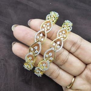 May include: Two gold-toned bangles with a floral design. The bangles feature a twisted pattern with clear, sparkling stones and small, leaf-like details. The design is set against a brown background.