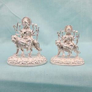 May include: Two silver statues of a deity riding a tiger. The deity has multiple arms and is adorned with intricate details. The statues are placed on ornate, circular bases. The background is a solid light blue color.