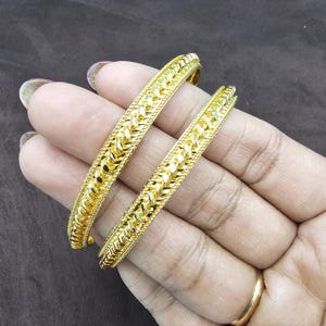 May include: Two gold-colored bangles with intricate woven patterns. The bangles are displayed on a hand, showcasing their detailed design and shiny finish. The jewelry is a bright, warm gold color.