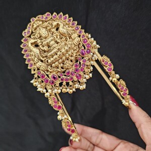May include: A gold hair ornament with a detailed design featuring a deity figure, surrounded by pink gemstones and pearl accents.