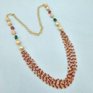 May include: Gold-toned necklace featuring a chain with alternating decorative beads and a cascade of small, burgundy beads. The necklace includes red, green, and clear accent beads. The design is intricate and elegant, suitable for special occasions.