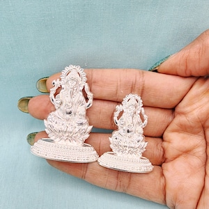 Pu&ograve; includere: Due statue color argento della divinit&agrave; ind&ugrave; Ganesha. Le statue sono finemente dettagliate, raffiguranti Ganesha seduto su una base di fiore di loto. Le statue sono tenute in una mano su uno sfondo azzurro.