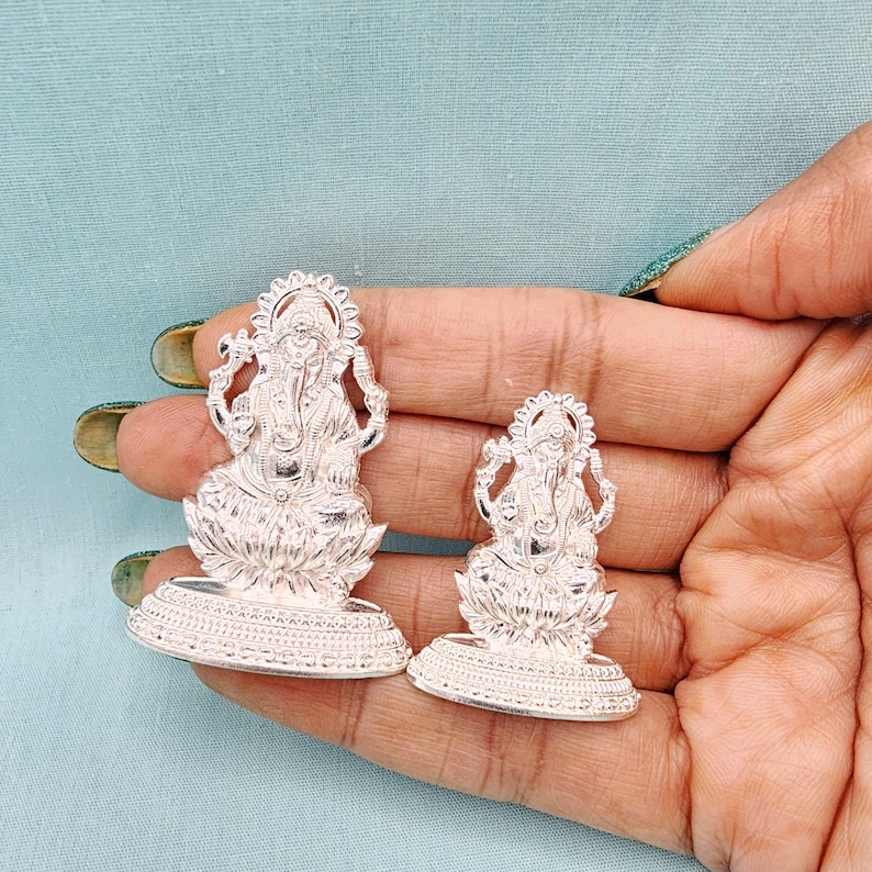 Pu&ograve; includere: Due statue color argento della divinit&agrave; ind&ugrave; Ganesha. Le statue sono finemente dettagliate, raffiguranti Ganesha seduto su una base di fiore di loto. Le statue sono tenute in una mano su uno sfondo azzurro.