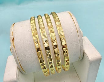 May include: Four gold bangles with intricate geometric patterns are displayed on a white stand against a light blue backdrop. The bangles have a repeating design of circles, lines, and squares, creating a textured appearance. The jewelry is presented in a studio setting.