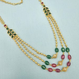 May include: A gold-colored necklace with three strands. Each strand features small, round beads and is accented with green and red oval-shaped gemstones. The necklace has leaf-shaped pendants with green gemstones.