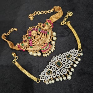 May include: Two gold choker necklaces with intricate designs. The top necklace features a peacock design with pink and green gemstones, while the bottom necklace has a geometric design with clear and green gemstones. Both necklaces have pearl accents.