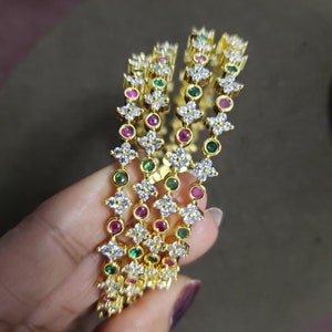 May include: Four gold bangles with white cubic zirconia stones and colorful accents. The bangles are stacked on top of each other and feature alternating green and pink stones.