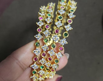 Puede incluir: Cuatro brazaletes de oro con piedras de circonita cúbica blancas y detalles coloridos. Los brazaletes están apilados uno encima del otro y presentan piedras verdes y rosas alternas.