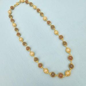 Puede incluir: Un collar dorado con cuentas esféricas alternas. Algunas cuentas son doradas, otras son de color rojo oscuro o dorado claro con un diseño de celosía. El collar tiene un cierre dorado.