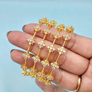 May include: Four gold-colored bangles with a floral design. Each bangle features a series of flower-shaped accents with clear, sparkling stones. The bangles are held in a hand, showcasing their intricate details and shine. The background is a light blue color.