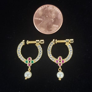 Puede incluir: Un par de pendientes de aro dorados con un diseño floral y una perla colgante. Los aros están incrustados con piedras preciosas claras y azules. Se incluye una moneda como referencia de tamaño.