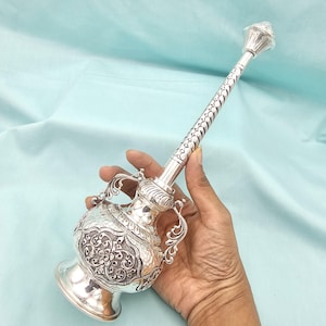 May include: A silver-colored, ornate vessel with a long, slender neck and a bulbous base. The vessel features intricate floral and geometric patterns, with two curved handles on the sides. The design is detailed and reflective, suggesting a decorative or ceremonial purpose.