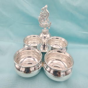 Puede incluir: Un servidor de condimentos plateado con cuatro cuencos redondos y un asa decorativa y ornamentada. El asa presenta un diseño detallado y los cuencos están dispuestos alrededor de un tallo central. El servidor está hecho de metal brillante.