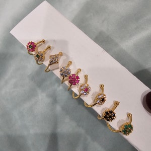 Può includere: Un set di otto anelli per il naso in oro con pietre preziose di diversi colori. Gli anelli sono disposti in fila su una superficie bianca. Le pietre preziose sono rosa, rosse, verdi, nere e bianche.