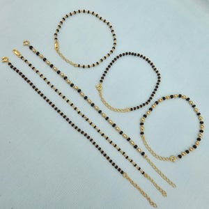 Op de afbeelding: Verschillende goudkleurige armbanden met zwarte kralen en gouden accenten worden getoond. De armbanden variëren in lengte en ontwerp, sommige met een kettingverlenging. De sieraden zijn gerangschikt op een lichtgekleurd oppervlak.