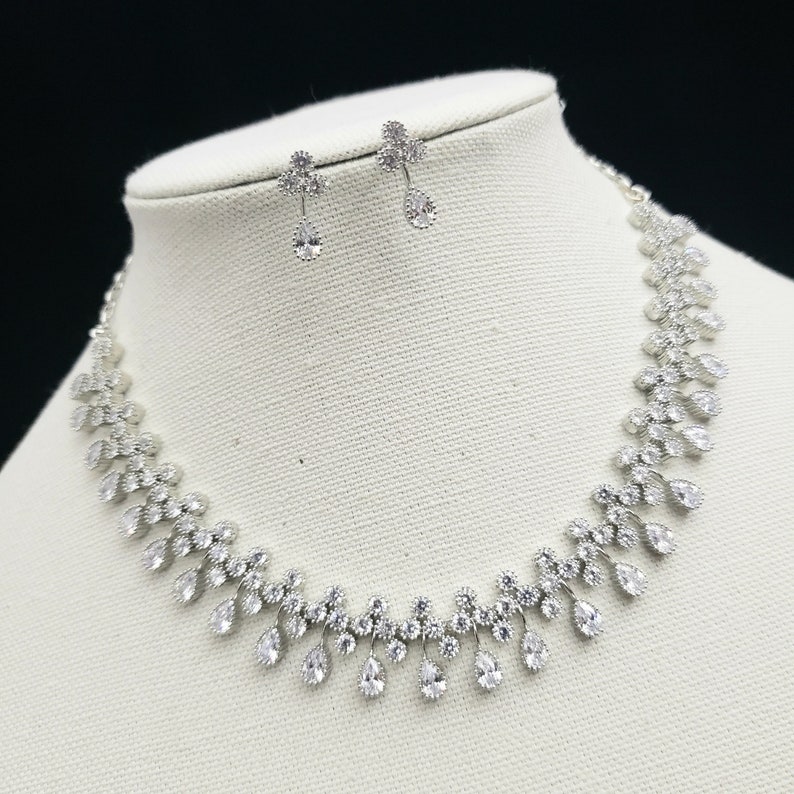 K&ouml;nnte beinhalten: Ein silbernes Halsketten- und Ohrring-Set mit einem Tropfen-Design. Die Halskette hat eine zarte Kette mit einer Reihe von tropfenf&ouml;rmigen Kristallen. Die Ohrringe sind ebenfalls tropfenf&ouml;rmig und h&auml;ngen an kleinen silbernen Ohrsteckern.