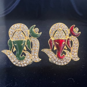 May include: Two gold-toned metal ornaments featuring the Hindu deity Ganesha. One ornament has a green Ganesha with red accents, and the other has a red Ganesha with green accents. Both ornaments are decorated with clear rhinestones.