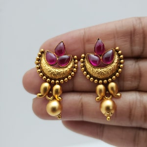 May include: A pair of gold-toned earrings featuring a crescent moon design with three teardrop-shaped, deep red gemstones. The earrings have a textured surface and small, round accents, with a dangling gold ball at the bottom.