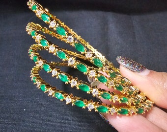 Puede incluir: Un conjunto de cuatro brazaletes de oro con piedras verdes y blancas. Los brazaletes están apilados uno encima del otro y están siendo sostenidos por una mano.