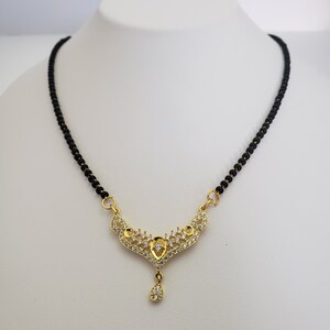 May include: A gold-colored necklace with a black beaded chain. The pendant features a decorative design with clear, sparkling stones. A teardrop-shaped stone hangs from the bottom of the pendant. The necklace is displayed on a white surface.