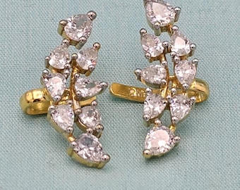 Pode incluir: Um par de brincos ear cuff dourados com um design curvo, adornados com múltiplas pedras transparentes em forma de pera. Cada peça apresenta um grupo de cristais facetados engastados numa estrutura metálica, criando um padrão de folha.