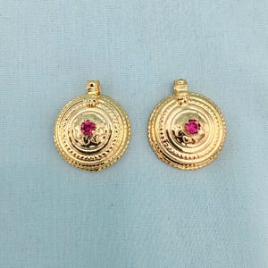 May include: Two gold-colored pendants with a circular design and a central red gemstone. The pendants have intricate detailing and a loop at the top for hanging. The background is a light blue color.