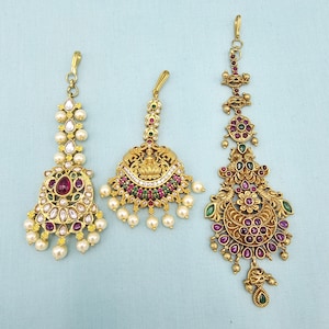 May include: Three ornate gold-toned headpieces with intricate designs. Each piece features a central pendant adorned with red, green, and white gemstones, and faux pearls. The headpieces are likely for special occasions.