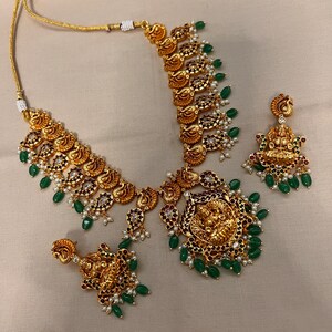 May include: A gold necklace and earrings set with a traditional Indian design. The necklace features a pendant with a deity figure, surrounded by paisley-shaped motifs and green and white beads. The earrings are similar in design, with a smaller pendant and matching beads.