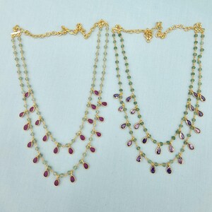 May include: Two gold necklaces with green beads and teardrop-shaped ruby and amethyst pendants. The necklaces are displayed on a light blue surface. The chains are delicate and the pendants are evenly spaced.