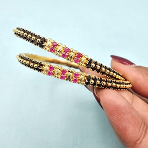 May include: Two gold bangles with alternating black and red gemstones. The bangles have a detailed design with small gold beads and decorative settings for the gemstones. The jewelry is displayed against a light blue background.