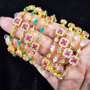May include: A set of six gold bangles with white and pink stones. The bangles are decorated with square-shaped designs and green stones.