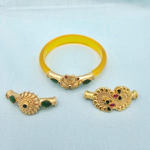 May include: A collection of gold-toned jewelry pieces, including a yellow bangle bracelet and two ornate clasps. The bracelet features gold accents with green and red gemstones. The clasps have intricate designs with similar gemstone embellishments.